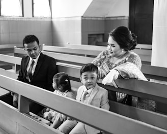 A family is seated on church pews. The scene includes a man in a suit, a woman in traditional attire, a young boy also dressed in a suit, and a girl in a dress. The woman and children seem engaged, with the man looking towards the girl.