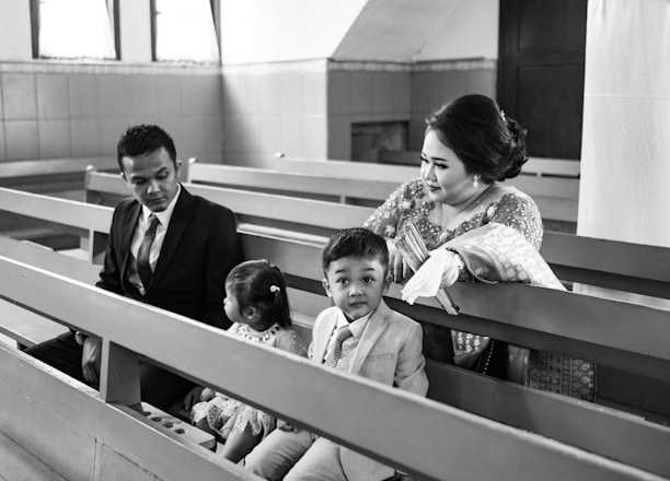 A family is seated on church pews. The scene includes a man in a suit, a woman in traditional attire, a young boy also dressed in a suit, and a girl in a dress. The woman and children seem engaged, with the man looking towards the girl.