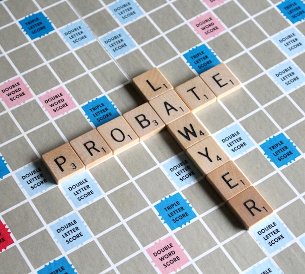 Wooden letter tiles spell out the words 'PROBATE' and 'LAWYER' on a Scrabble board. The board features a grid with colored squares marked with different point values such as 'Double Word Score' and 'Triple Letter Score'.