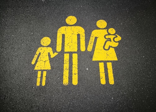 Yellow painted silhouettes on a dark gray asphalt background depicting a family consisting of two adults, one child, and a baby.