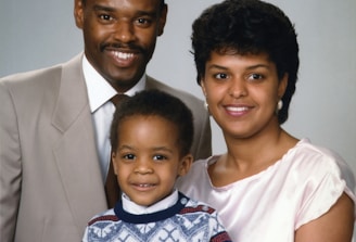 A family portrait featuring three people, consisting of two adults and a child. The man is wearing a beige suit and a tie, while the woman is dressed in a light pink top. The child is standing in front, wearing a patterned sweater. All appear to be smiling and are posed closely together in front of a neutral background.