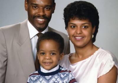 A family portrait featuring three people, consisting of two adults and a child. The man is wearing a beige suit and a tie, while the woman is dressed in a light pink top. The child is standing in front, wearing a patterned sweater. All appear to be smiling and are posed closely together in front of a neutral background.