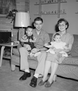 A family of four is seated on a couch in a living room. The father holds a young boy in his lap, while the mother holds an infant girl. The room features a fireplace with a decorative clock on the mantel, a mid-century style lamp, a side table with a radio, and shelves with various figurines and decorations. The family appears happy and well-dressed, with the father and son in button-up shirts and slacks, and the mother wearing glasses, a blouse, and a skirt.