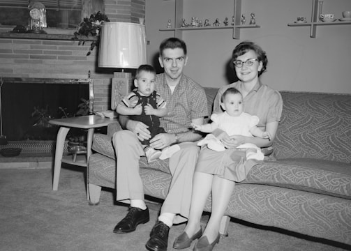 A family of four is seated on a couch in a living room. The father holds a young boy in his lap, while the mother holds an infant girl. The room features a fireplace with a decorative clock on the mantel, a mid-century style lamp, a side table with a radio, and shelves with various figurines and decorations. The family appears happy and well-dressed, with the father and son in button-up shirts and slacks, and the mother wearing glasses, a blouse, and a skirt.