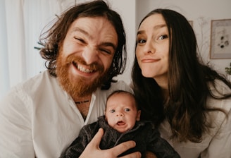 A cheerful family portrait featuring a bearded man with long hair smiling widely, a woman with straight long hair making a playful expression, and a baby with a curious look. They are all dressed in casual attire with light-colored backgrounds suggesting an indoor setting.