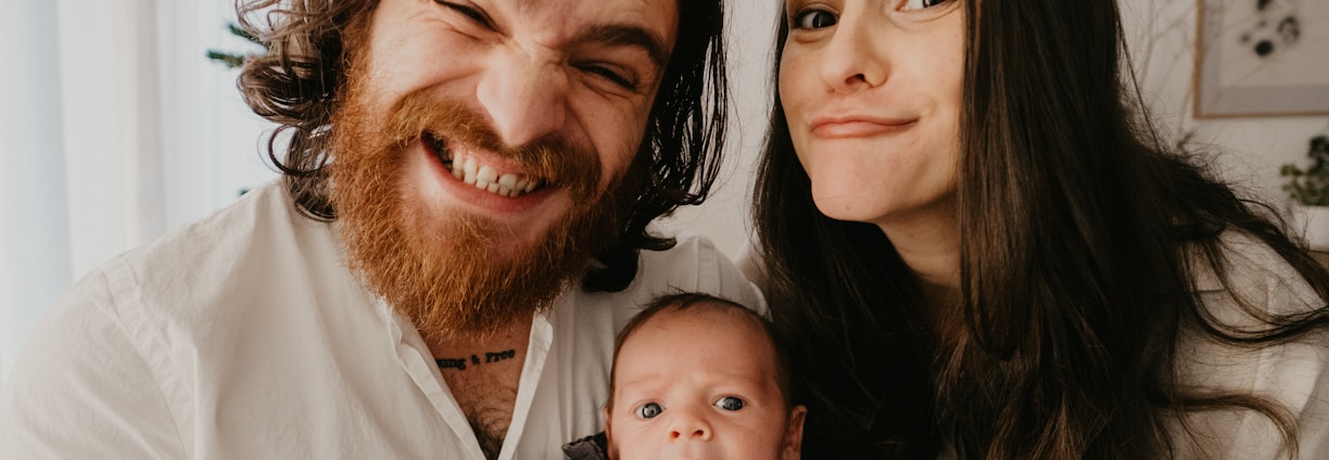 A cheerful family portrait featuring a bearded man with long hair smiling widely, a woman with straight long hair making a playful expression, and a baby with a curious look. They are all dressed in casual attire with light-colored backgrounds suggesting an indoor setting.