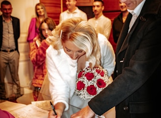 A woman in a white dress is signing a document on a table while holding a bouquet of red and white flowers. Several people stand around her, including a person pointing at the document. The room has wooden paneling and a warm ambiance.