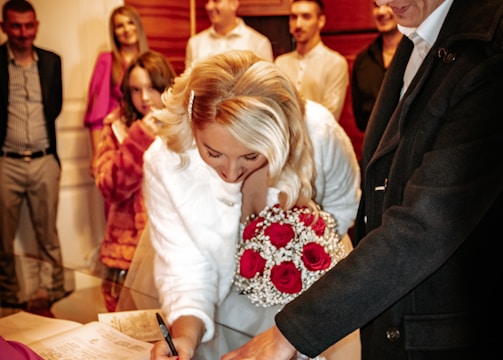 A woman in a white dress is signing a document on a table while holding a bouquet of red and white flowers. Several people stand around her, including a person pointing at the document. The room has wooden paneling and a warm ambiance.