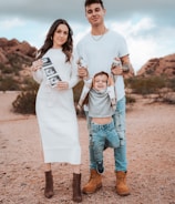 A family standing outdoors in a desert-like setting with a rocky backdrop. The woman is wearing a white dress and holding ultrasound images, while the man in a white shirt and ripped jeans is holding a child by the arms, playfully suspending them in front.