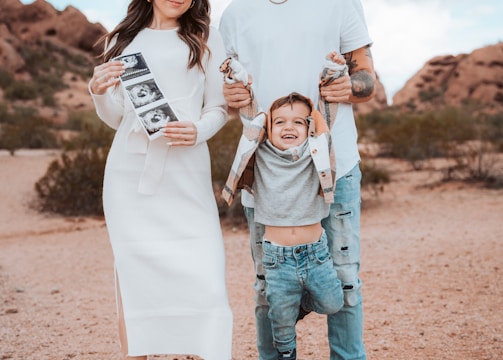 A family standing outdoors in a desert-like setting with a rocky backdrop. The woman is wearing a white dress and holding ultrasound images, while the man in a white shirt and ripped jeans is holding a child by the arms, playfully suspending them in front.