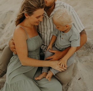 A family sitting together on a sandy beach, with an adult man embracing a woman and child. The woman is wearing a long, flowing light green dress, while the child is dressed in a gray outfit. The setting seems peaceful and intimate, with a warm and relaxed atmosphere.