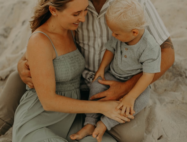 A family sitting together on a sandy beach, with an adult man embracing a woman and child. The woman is wearing a long, flowing light green dress, while the child is dressed in a gray outfit. The setting seems peaceful and intimate, with a warm and relaxed atmosphere.