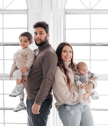 A family portrait featuring two adults and two children standing in front of large, bright windows. The father holds a toddler, while the mother holds a baby. They are all dressed in cozy, neutral-toned sweaters and jeans.