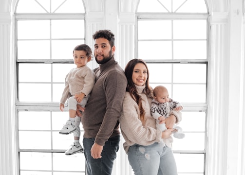 A family portrait featuring two adults and two children standing in front of large, bright windows. The father holds a toddler, while the mother holds a baby. They are all dressed in cozy, neutral-toned sweaters and jeans.