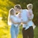 A family is standing in a sunlit field. The adults are sharing a kiss while holding a toddler and a baby. The sunlight creates a warm glow around them, highlighting their expressions. The father's head is turned towards the mother, and they are surrounded by green foliage.