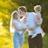 A family is standing in a sunlit field. The adults are sharing a kiss while holding a toddler and a baby. The sunlight creates a warm glow around them, highlighting their expressions. The father's head is turned towards the mother, and they are surrounded by green foliage.