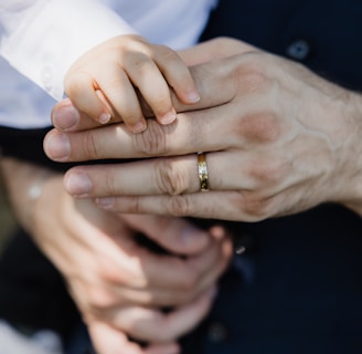 A close-up of a small child's hand gently placed on top of an adult's hand, which is wearing a gold band. The adult is dressed in a dark suit with a white shirt, and the child's sleeve is also white. The background is softly blurred, drawing focus to the hands.