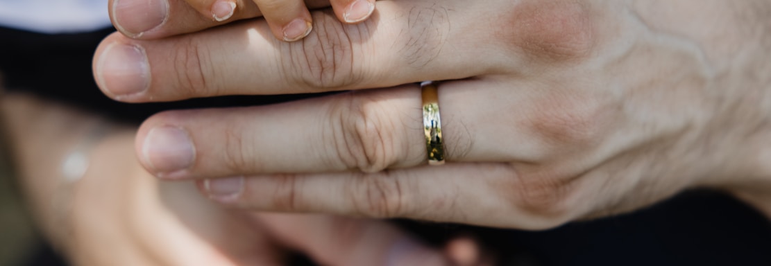 A close-up of a small child's hand gently placed on top of an adult's hand, which is wearing a gold band. The adult is dressed in a dark suit with a white shirt, and the child's sleeve is also white. The background is softly blurred, drawing focus to the hands.