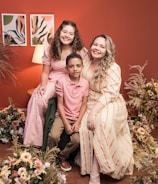 A joyful family portrait featuring three people sitting together. They are surrounded by floral arrangements. The background includes a warm red wall with two art pieces and a lit lamp, creating a cozy atmosphere.