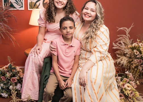 A joyful family portrait featuring three people sitting together. They are surrounded by floral arrangements. The background includes a warm red wall with two art pieces and a lit lamp, creating a cozy atmosphere.