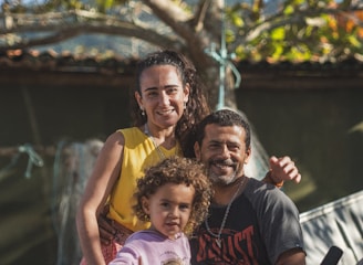 A joyful family portrait with a smiling man, woman, and child in an outdoor setting. The man and woman are sitting close together, while the child is in the foreground, making a playful gesture. The background features a tree with green and yellow leaves, suggesting a pleasant sunny day.