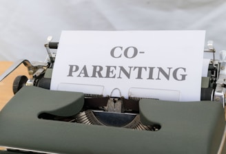 A close-up view of a vintage typewriter displaying a piece of paper with the word 'CO-PARENTING' typed in bold letters. The typewriter has a dark green cover and is placed on a light wooden desk with a neutral background.