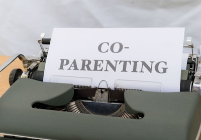 A close-up view of a vintage typewriter displaying a piece of paper with the word 'CO-PARENTING' typed in bold letters. The typewriter has a dark green cover and is placed on a light wooden desk with a neutral background.