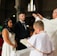 An indoor wedding ceremony with a bride in a white dress and a groom in a dark suit standing before a priest. The setting is a church with arched windows and brick walls. A child in a white robe stands nearby holding a book.