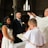 An indoor wedding ceremony with a bride in a white dress and a groom in a dark suit standing before a priest. The setting is a church with arched windows and brick walls. A child in a white robe stands nearby holding a book.